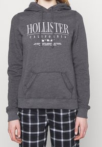 Grå hoodie med en framficka, dragsko huva och vit "Hollister California" logotyp. Bärs över svart- och vitrutiga byxor.
