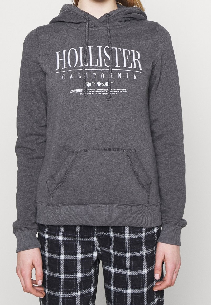 Grå hoodie med en framficka, dragsko huva och vit "Hollister California" logotyp. Bärs över svart- och vitrutiga byxor.