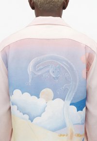 Άτομο που φορά ένα πουκάμισο απαλού ροζ χρώματος με ένα διαφανές σχέδιο δράκου-γάτας πάνω από τα σύννεφα και έναν ήλιο στην πλάτη.