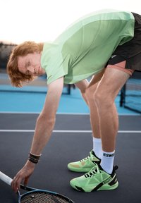 Camisa deportiva verde y pantalones cortos negros; calzado K-Swiss verde menta. El jugador se está agachando para recoger una raqueta de tenis en una cancha.