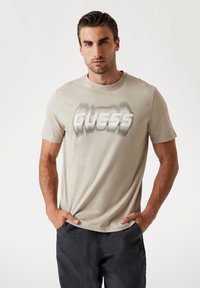 Lühikeste varrukatega beež puuvillane T-särk, mille esiküljel on julge, tuhmunud "GUESS" graafika, kantud tumeda tekstuuriga lühikeste pükstega.