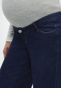 Κοντινό πλάνο εγκύου κοιλιάς με γκρι τοπ και σκούρο μπλε παντελόνι εγκυμοσύνης με ξεκουμπωμένη τη μέση για άνεση.