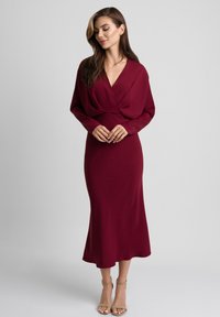 Burgundowa sukienka wrap z długim rękawem, dekoltem w kształcie V, dopasowaną talią i swobodną spódnicą. Wykonana z gładkiego materiału, z delikatnymi detalami w drapowaniach.