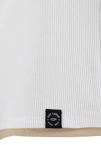 Tissu blanc à côtes avec un ourlet beige. Présente une étiquette noire indiquant "Key Largo" et "Depuis 1994" dans le coin inférieur gauche.