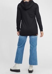 Černá mikina s klokankou, kapucí se stahovací šňůrkou a hnědou koženou visačkou. V kombinaci s světle modrými širokými džíny a bílými kotníkovými botami.