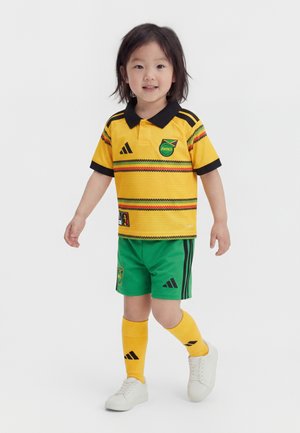 Niño pequeño con uniforme de fútbol de Jamaica en amarillo y verde, de pie con un pie ligeramente adelantado sobre un fondo blanco liso.