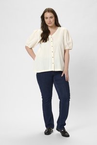 Blouse ample crème avec des manches bouffantes, cinq boutons marron sur le devant, assortie à un jean ajusté bleu foncé et des chaussures noires.