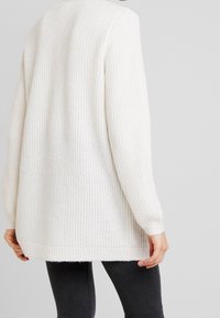 Biały sweter dzianinowy o luźnym kroju, z prążkowaną teksturą i długimi rękawami. Dolna część jest nieco dłuższa z tyłu.