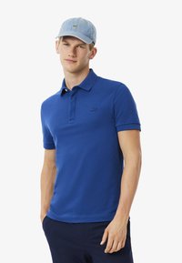 Blå polo skjorta med mjuk textur, korta ärmar och krage. Har en diskret logotyp på vänster bröst. Matchas med en ljusblå keps.