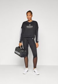 Nasmejana žena stoji u sportskom odijelu: crna dukserica i tajice, držeći crnu Adidas torbu, nasuprot neutralnoj pozadini.