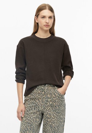 Vrouw met lang haar die een zwarte trui en luipaardprintbroek draagt, staat met één hand in haar zak tegen een witte achtergrond.