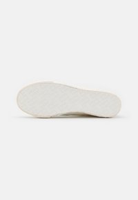 Sålene af en hvid sneakers med et geometrisk rektangulært mønster præget på gummiydersålen, præsenteret mod en ensfarvet hvid baggrund.
