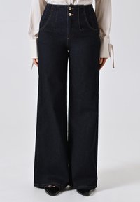 Brede donkere denim jeans hebben een hoge taille met twee voorknopen, contrasterende stiksels en zijzakken. Gladde textuur en volle lengte.