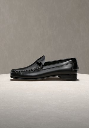 Mocassin pour homme en cuir noir avec surpiqûres, posé sur une surface beige devant un fond gris.