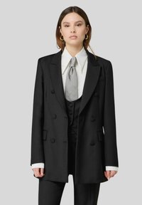 Blazer nero doppiopetto con bottoni prominenti, revers a scapolo e una vestibilità strutturata. Presenta una camicia bianca e una cravatta grigia.