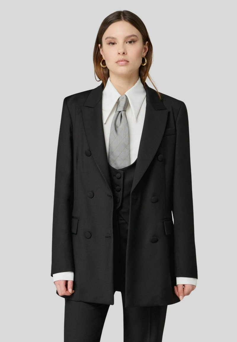 Blazer nero doppiopetto con bottoni prominenti, revers a scapolo e una vestibilità strutturata. Presenta una camicia bianca e una cravatta grigia.