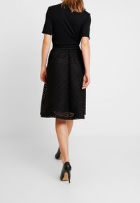 Schwarzes kurzärmeliges Oberteil kombiniert mit einem strukturierten schwarzen Midirock mit halbtransparentem Muster. Dazu schwarze High-Heel-Schuhe getragen.