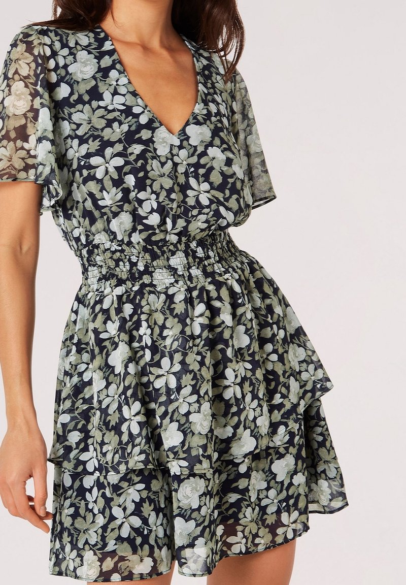 Robe fleurie à col en V, manches courtes et taille smockée. Caractérisée par des jupes superposées en bleu marine foncé avec des motifs floraux verts. Tissu transparent.