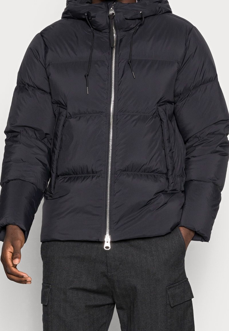 Osoba ubrana w czarną kurtkę puchową z zamkiem błyskawicznym z przodu i kapturem, w połączeniu z ciemnymi spodniami cargo, z dłońmi częściowo w kieszeniach.