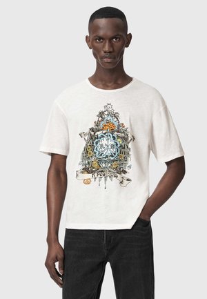 Mann trägt weißes T-Shirt mit buntem Blumen- und abstraktem Grafikmotiv in der Mitte vorne, kombiniert mit schwarzen Jeans, steht vor hellem Hintergrund.