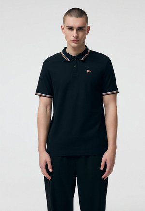 Młody mężczyzna z krótkimi włosami, ubrany w czarną koszulkę polo z pomarańczowo-białym paskowanym kołnierzem i brzegami rękawów, stojący na jasnym, jednolitym tle.