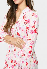 Robe fleurie avec un fond clair, ornée de fleurs rouges et roses. Manches courtes, décolleté en V, avec une taille froncée et une jupe fluide.