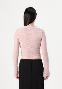 Femme aux longs cheveux noirs portant un haut rose en dentelle ajusté à manches longues et un pantalon noir taille haute, debout de dos sur fond blanc.