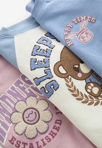 Tre tröjor staplade: ljusblå, vit och lavendel. Med broderade mönster av en björn, en leende blomma och textdetaljer.