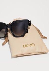 Ochelari de soare negri în formă de ochi de pisică, cu lentile degradate, asortați cu un pouch bej ce prezintă branding-ul auriu "LIU-JO".