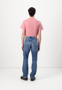 Camiseta rosa claro de manga corta, jeans de mezclilla azules con corte recto y bolsillos traseros, combinados con zapatos negros tipo slip-on, vistos desde atrás.