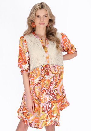 Donna con lunghi capelli biondi che indossa un vestito floreale arancione, rosa e giallo con un gilet in pizzo beige, posando con una mano sui fianchi.