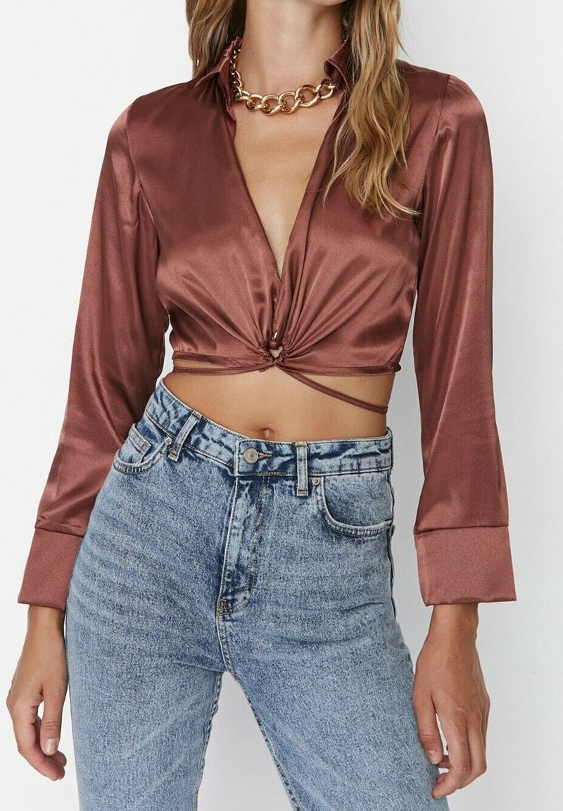 Femme portant un crop top en satin couleur rouille avec des manches longues, un nœud à l'avant, et un jean taille haute bleu clair, accessoirisée d'un collier chaîne dorée épais.
