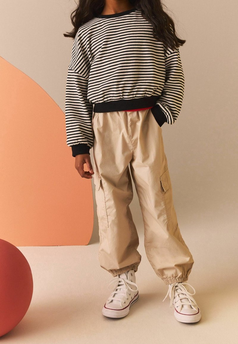 Enfant portant un t-shirt à manches longues rayé noir et blanc, un pantalon cargo beige, et des baskets montantes blanches sur un fond neutre.