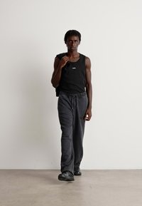 Black tank top with a small logo, gray sweatpants, and black sneakers, worn by a person walking against a light background.