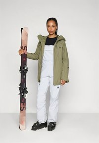 Suusariietus, mis sisaldab tumerohelist veekindlat jopet ja helehallid üleülikonda, mustad suusasaapad ja käes paar suuski.