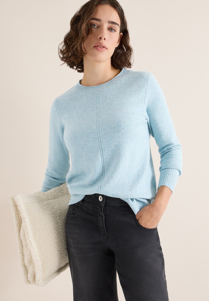 Hellblauer Pullover aus weichem Material mit rundem Ausschnitt, gerippten Bündchen und einem dezenten Nahtdetail. Kombiniert mit schwarzen Jeans und einem cremefarbenen Accessoire.