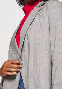 Grey plaid blazer with light orange and pink lines. Features a single button closure and notched lapels, set against a red turtleneck.