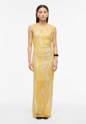 Femme portant une robe sans manches, longue jusqu'au sol, à motifs ondulés jaunes et beiges, un bracelet noir et des boucles d'oreilles anneaux argentés, debout sur un fond blanc.