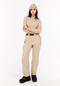 Beige top met lange mouwen en een ritssluiting bij de hals, gecombineerd met beige broek met elastische taille. Zwarte schoenen met bandjes maken de outfit compleet.