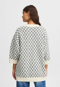 Weißer Strickpullover mit einem schwarzen Rautenmuster, gerippten Bündchen und Saum. Lockerer Schnitt und mittellange Ärmel. Über blauen Denim-Jeans getragen.