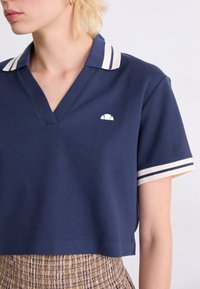 Navy cropped polo shirt with a V-neck and white striped collar, featuring a small white logo on the left chest. Classic cotton texture.