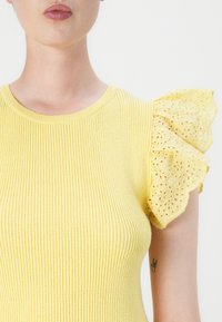 Gul ribbet topp med korte volangermer som har hullbroderi. Utringningen er rund, og stoffet har en myk tekstur.