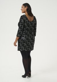 Robe noire à sequins avec des manches trois-quarts, décolleté plongeant, forme ajustée et motif floral. Associée avec des collants noirs et des talons.