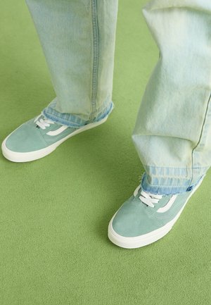 Jasnozielone sneakersy z białymi akcentami, wykonane z zamszu, na płaskiej podeszwie. Noszone z jasnymi dżinsami, lekko postrzępionymi na nogawkach, na zielonej powierzchni.