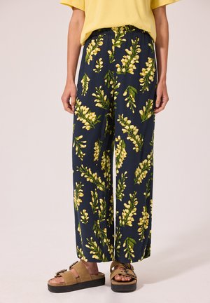 Pantalon large bleu marine à imprimé floral jaune, porté avec des sandales marron clair et un top jaune à manches courtes, montré de la taille jusqu'aux pieds.