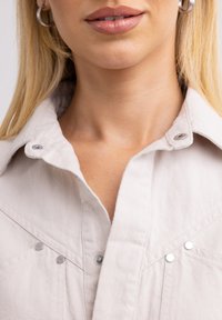 Kreemivärvi denimpoolt, millel on nupud, järgneva terava kraega ja ette dekoratiivsete metallaktsentidega. Sile tekstuur.