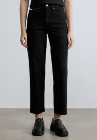 Μαύροι τζιν ψηλόμεσοι από τζιν ύφασμα, με ίσια γραμμή, θηλιές για ζώνη και μπροστινές τσέπες. Το μοντέλο φοράει μαύρα παπούτσια slip-on.