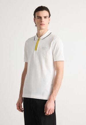 Polo bianco con rifiniture nere e una cerniera gialla. Presenta un piccolo logo azzurro chiaro sul lato sinistro del petto. Maniche corte, materiale in cotone.