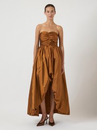 Strapless brown satin gown with gathered bodice, full skirt with asymmetrical hem, and side slit. Styled with pointed brown heels.