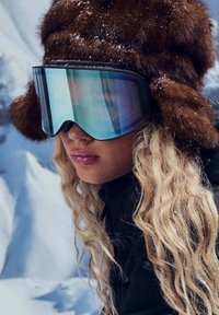 Brauner Pelzmütze mit Ohrenschützern, oben mit Schneeflocken. Große reflektierende Skibrille mit blauem Verlaufsfilter. Welliges blondes Haar sichtbar.